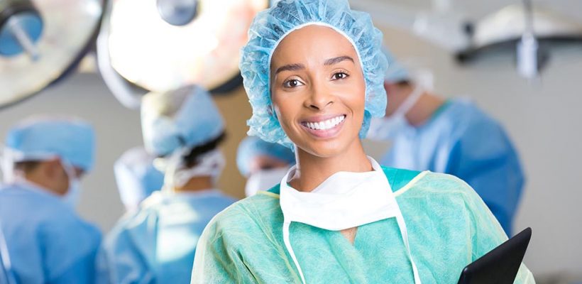 Pretty young African American female surgeon prepares for surgery. She is wearing surgical gown, surgical mask and hair net. She is holding a digital tablet. a team of surgeons are operating in the background.